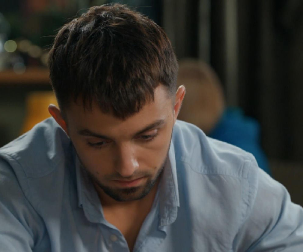 Młody mężczyzna, Mirek (Oliwier Kozłowski) z serialu Barwy szczęścia, spogląda w dół z zatroskaną miną, w jasnoniebieskiej koszuli. Jest to zdjęcie z odcinka 3318, gdzie Mirek zmaga się z konsekwencjami swojego romansu i powrotem na boisko, o czym przeczytasz więcej na Super Seriale.