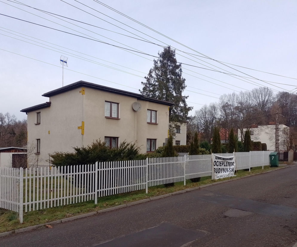 Piętrowy dom typu kostka za białym płotem, na którym wisi ogłoszenie o ocieplaniu budynków. Więcej na Muratordom.