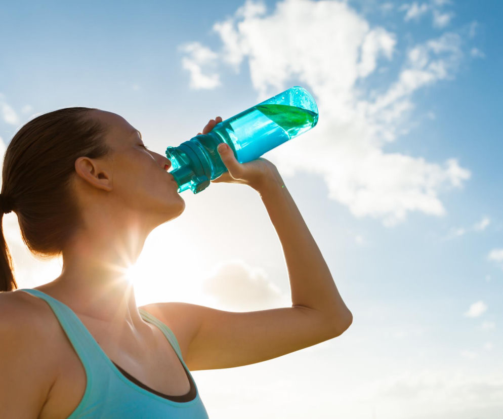 Kobieta w błękitnym topie pije wodę z przezroczystej, niebieskiej butelki, trzymając ją wysoko. Twarz jest widoczna z profilu, a w tle rozciąga się bezchmurne niebo. Słońce świeci zza postaci, tworząc promienisty blask. O tym, jak prawidłowo dbać o butelki wielorazowe, dowiesz się na Poradnik Zdrowie.