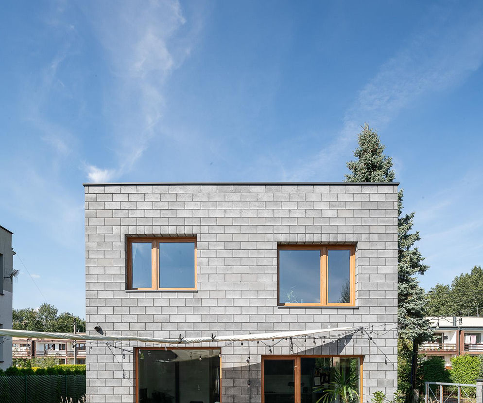 Dwukondygnacyjny Dom w Ochojcu projektu Jojko+Nawrocki Architekci, z szarą elewacją z pustaków betonowych i drewnianymi oknami, otoczony zielonym ogrodem z tarasem z desek. Ten dom to przykład przemyślanej Architektury Murator Plus.