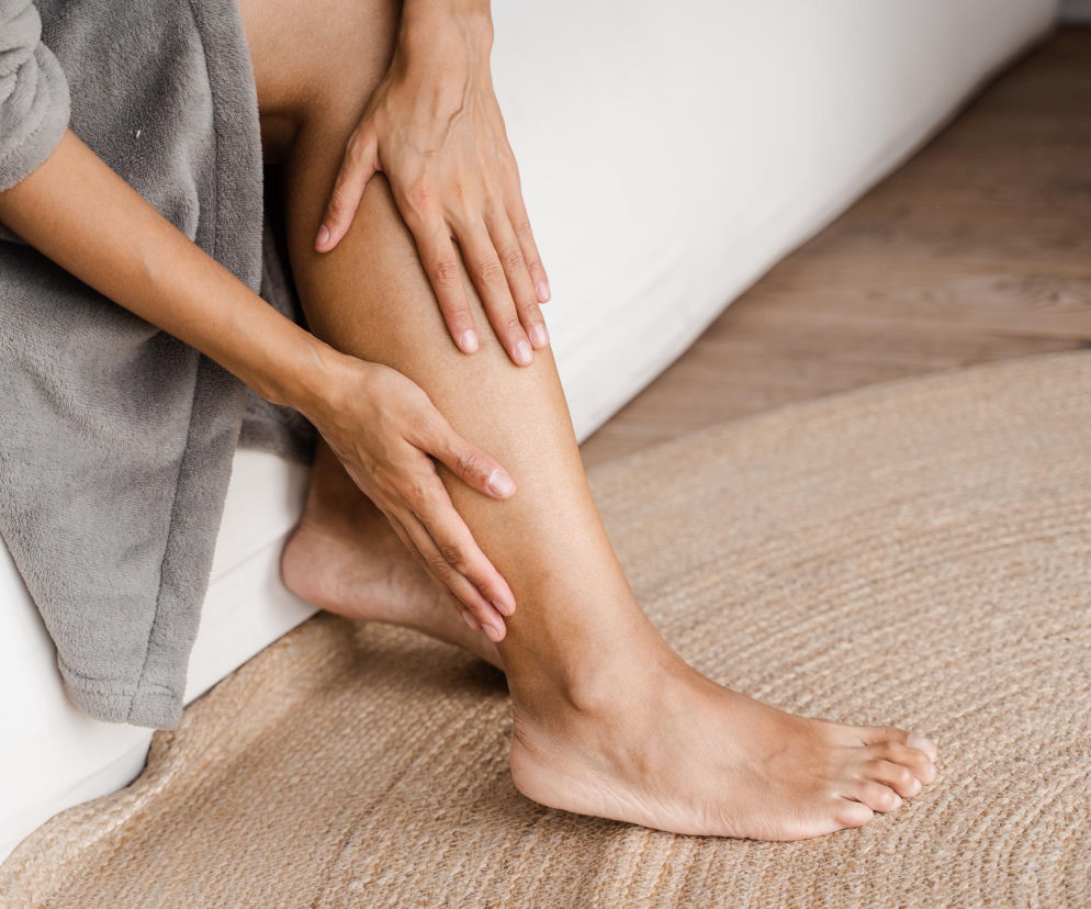 Kobieta w szlafroku masuje bolące łydki i stopy na jasnym dywanie, co może wskazywać na problem z bólem stóp i pleców. Na Poradniku Zdrowie dowiesz się, jak łatwo zaradzić takim dolegliwościom, by pozbyć się ich na dobre.