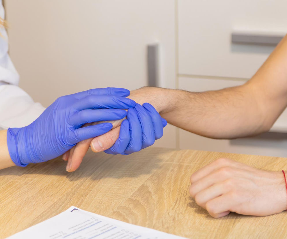 Lekarka w niebieskich rękawiczkach bada dłoń pacjenta, sprawdzając objawy akromegalii. Na drewnianym biurku leży dokumentacja medyczna. Akromegalię można wykryć szybciej dzięki AI, o czym przeczytasz na Poradnik Zdrowie.