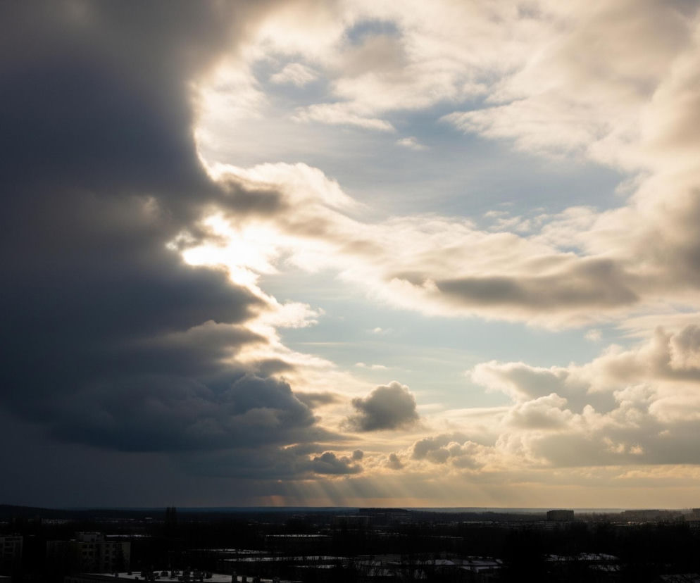 Niebo wypełniają chmury o różnej jasności i zabarwieniu, od ciemnoszarych po biało-złote. W centralnej części obrazu widoczne jest przejaśnienie w chmurach, przez które widać jaśniejsze, błękitne niebo. Promienie słońca przebijają się przez niżej położone chmury, tworząc smugi światła o złotawym odcieniu, które rozciągają się w kierunku horyzontu. Poniżej, w ciemnej, słabo oświetlonej części obrazu, rozciąga się miejski krajobraz z widocznymi budynkami i drzewami, zlewającymi się z ciemnym tłem.