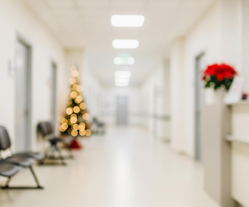 Rozmazany obraz przedstawia długi korytarz z kremowymi ścianami i jasną podłogą, rozciągający się od pierwszego planu w głąb perspektywy. Po lewej stronie widoczne są trzy czarne, połączone krzesła, a za nimi, w oddali, zamglona choinka ze świecącymi, okrągłymi światłami w ciepłym, żółtym kolorze. Po prawej stronie, na pierwszym planie, znajduje się fragment blatu lub lady recepcyjnej, na której stoi wazon z jaskrawoczerwonymi kwiatami i ciemne obiekty przypominające monitory lub tablety.