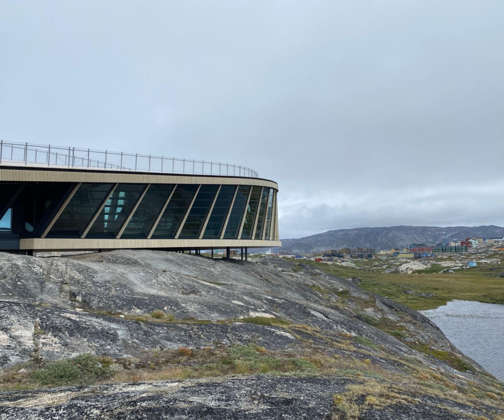 Centrum turystyczne Ilulissat Icefjord na Grenlandii autorstwa Dorte Mandrup, z charakterystycznym dachem z barierką, wykończone drewnem i szkłem, uniesione na palach nad skalistym terenem. W tle widać jezioro oraz zabudowania. Więcej o tej architekturze na Architektura Murator Plus.
