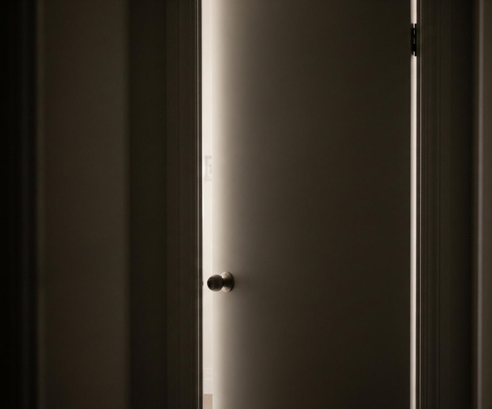 Szczelina otwartych drzwi, wypełniona jasnym, pionowym pasmem światła, dzieli obraz na dwie części. Po lewej stronie dominuje ciemna, jednolita płaszczyzna ściany, częściowo oświetlona delikatną poświatą. Z prawej strony widać fragment drzwi w odcieniach szarości z okrągłą, metalową klamką na wysokości około jednej trzeciej wysokości od dołu; klamka odbija światło, tworząc refleksy. Górny prawy róg ukazuje zawias drzwi oraz cienie na framudze, podkreślające trójwymiarowość ujęcia.