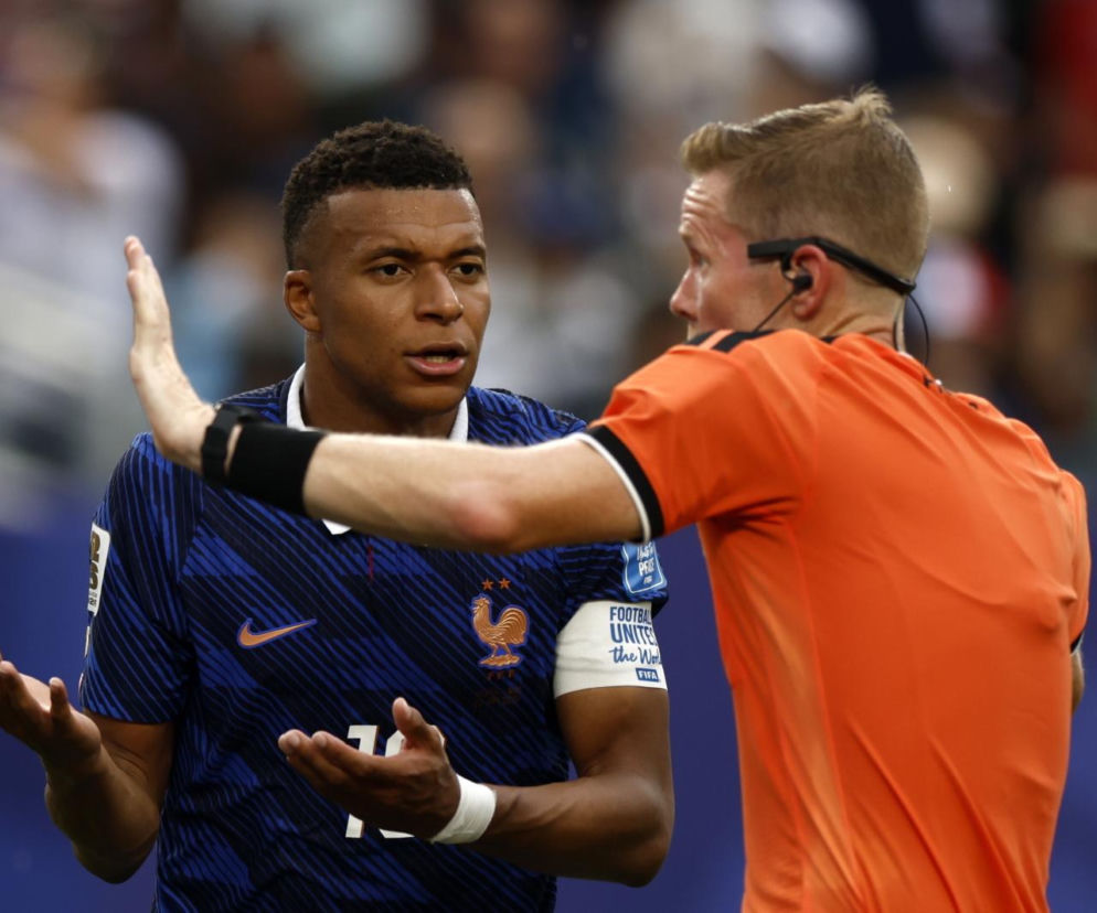 Piłkarz Kylian Mbappe w granatowej koszulce reprezentacji Francji, rozmawiający z arbitrem w pomarańczowej koszulce podczas meczu mistrzostw świata. Arbiter, z dłonią uniesioną w geście zatrzymania, rozmawia z zawodnikiem. Więcej o przerwanych meczach przeczytasz na naszym portalu.