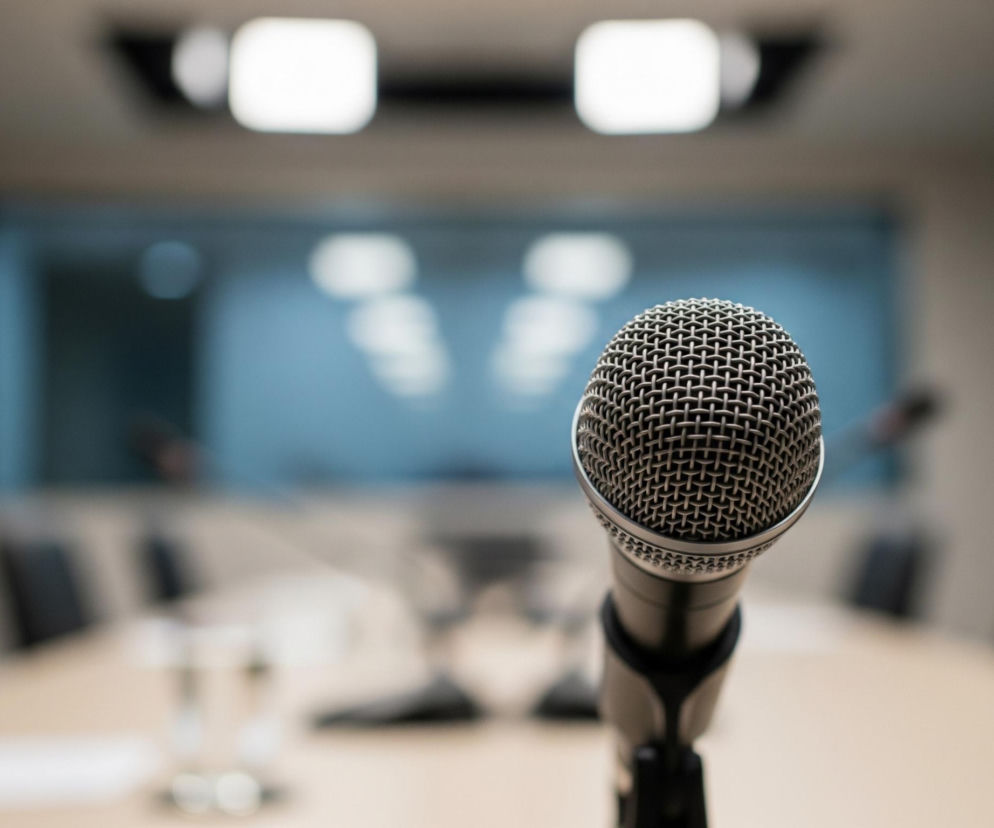 Centralnie umiejscowiony jest srebrny mikrofon na czarnym statywie, z siateczkową osłoną główki, która jest ostra i szczegółowa. Tło, rozmyte, przedstawia wnętrze sali konferencyjnej, z długim, jasnym stołem i rzędami ciemnych krzeseł po obu stronach. Na stole widoczne są niewyraźne zarysy szklanek i dodatkowych mikrofonów, a w oddali duża, jasna ściana z niebieskawymi panelami, nad którymi widać podłużne, prostokątne światła sufitowe.