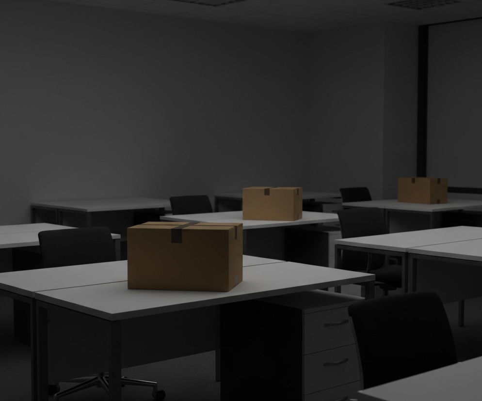W przestronnym pomieszczeniu biurowym dominują rzędy biurek w jasnoszarym kolorze z ciemnymi, metalowymi nogami, ustawione na ciemnym dywanie. Na pierwszym planie, w centrum, znajduje się jedno z tych biurek, a na nim brązowe kartonowe pudełko z czarną taśmą klejącą na wieczku, tworzącą kształt krzyża. W tle widać więcej podobnych biurek, na niektórych z nich również stoją identyczne kartonowe pudełka. Pomieszczenie oświetlone jest słabo, głównie światłem padającym z góry, co tworzy długie cienie i ponury nastrój.