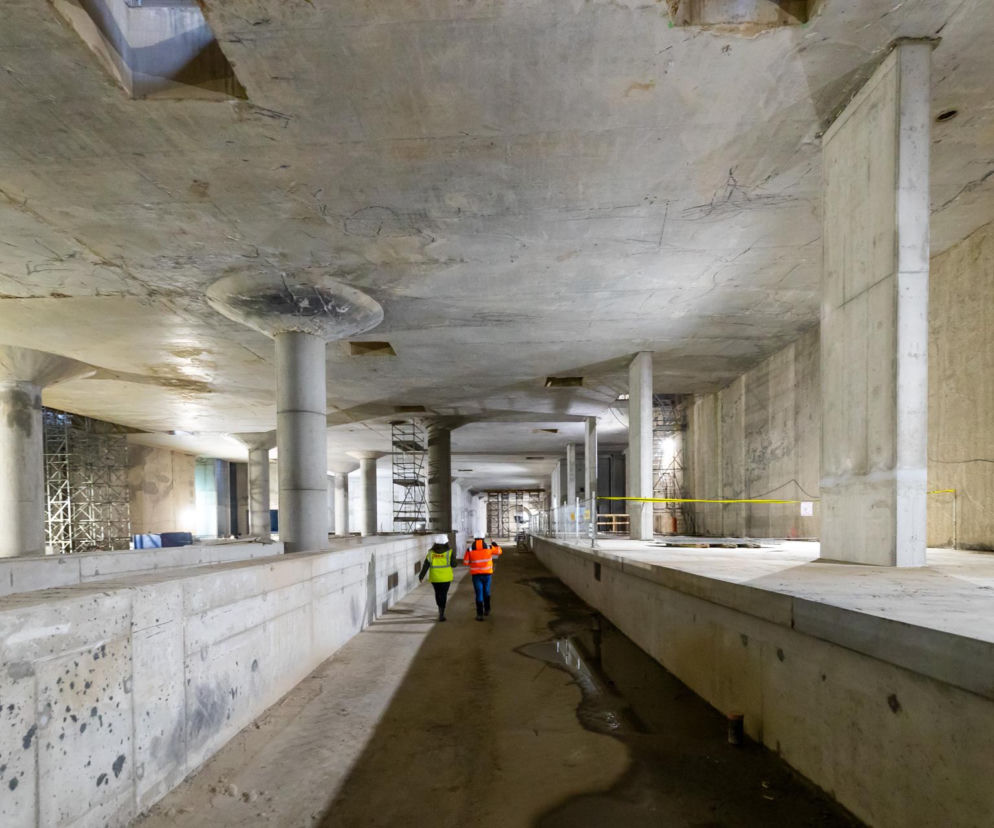 Długi betonowy tunel w trakcie budowy. Dwie osoby w kaskach i kamizelkach odblaskowych idą w głąb tunelu. Tunel średnicowy w Łodzi to największa inwestycja kolejowa w Polsce, o której przeczytasz na portalu Architektura Murator Plus.