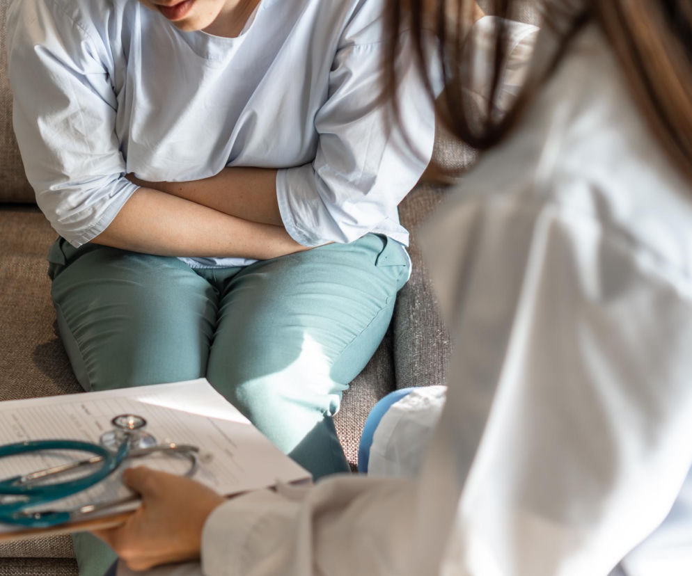 Kobieta z bólami brzucha siedzi na kanapie podczas wizyty u ginekologa. Lekarz trzyma w dłoniach ankietę medyczną i stetoskop. O endometriozie, jej objawach i sposobach leczenia, dowiesz się więcej na portalu Poradnik Zdrowie.