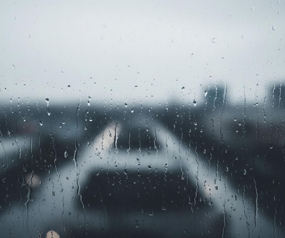 Szyba pokryta jest licznymi kroplami deszczu o różnych rozmiarach, niektóre tworzą smugi spływające w dół. Za zmoczonym szkłem widać rozmyty pejzaż miejski utrzymany w odcieniach szarości i ciemnego błękitu, z niewyraźnymi zarysami budynków i ulic. W tle widoczne są również pojedyncze, rozmyte światła, które przyjmują kształt okrągłych, jasnych plam. Ogólna atmosfera obrazu jest melancholijna i spokojna.