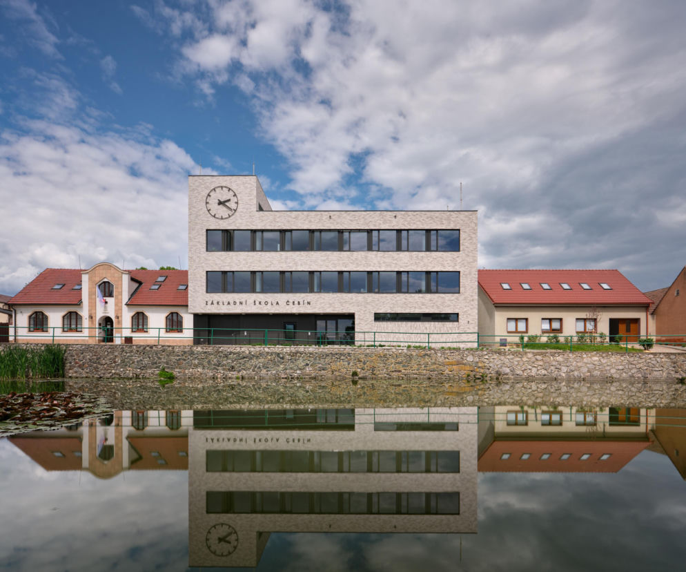 Szkoła w Čebínie z jasną elewacją i zegarem odbija się w wodzie. O tej przebudowie przeczytasz w serwisie Architektura.