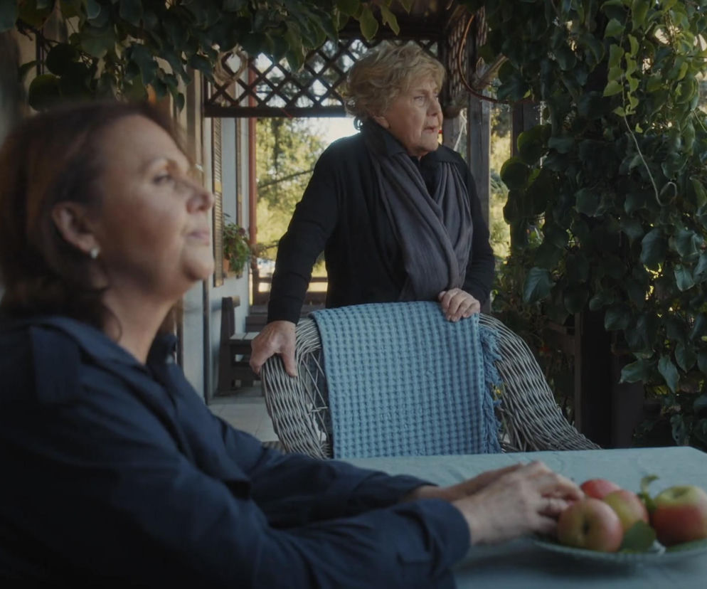 Kobiety, Barbara (Teresa Lipowska) i Marysia (Małgorzata Pieńkowska), na werandzie domu w Grabinie. Barbara stoi za wiklinowym krzesłem z niebieskim kocem, Marysia siedzi przy stole z jabłkami. Scena z serialu M jak miłość, o którym więcej przeczytasz na Super Seriale.