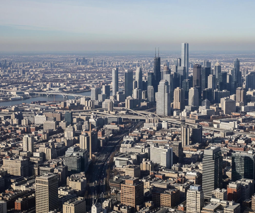 Rozległa panorama miasta z lotu ptaka ukazuje gęstą zabudowę, z licznymi wieżowcami dominującymi w centrum, oraz rozciągającą się na horyzoncie siatkę miejską. W centrum kadru wznosi się skupisko wysokich, nowoczesnych drapaczy chmur o szarych i granatowych fasadach, z jednym wyraźnie wyższym, smukłym budynkiem o błękitnych odbiciach w górnej części oraz ciemnym wieżowcem z dwoma antenami na szczycie. W lewej części obrazu widoczna jest rzeka przepływająca przez miasto, nad którą rozciągają się liczne mosty, w tym duży most łukowy i kilka estakad drogowych. Reszta obrazu to mozaika niższych budynków o zróżnicowanej architekturze, z przeważającymi odcieniami brązu, beżu i szarości, tworzących rozległy miejski krajobraz pod jasnym, nieco zamglonym niebem.