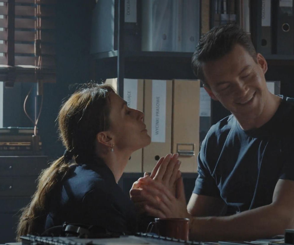 Martyna (Magdalena Turczeniwicz) klęcząca przed Jakubem (Krzysztof Kwiatkowski) z rękami złożonymi w błagalnym geście w biurze detektywistycznym, w tle biurowe segregatory. Mężczyzna uśmiecha się lekko, zaskoczony jej prośbą. Więcej o ich relacji przeczytasz na portalu Super Seriale.