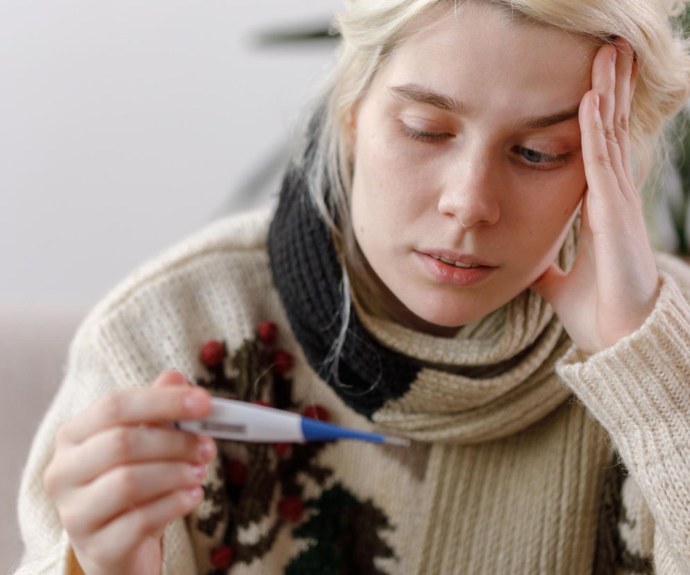 Młoda kobieta z jasnymi włosami, w ciepłym swetrze i szaliku, trzymająca termometr i mierząca temperaturę, z dłonią opartą na skroni, co sugeruje złe samopoczucie. Zdjęcie ilustruje zdradliwe objawy przeziębienia, które mogą być początkiem poważniejszych chorób. O tym, jak rozpoznać objawy, dowiesz się na Poradnik Zdrowie.