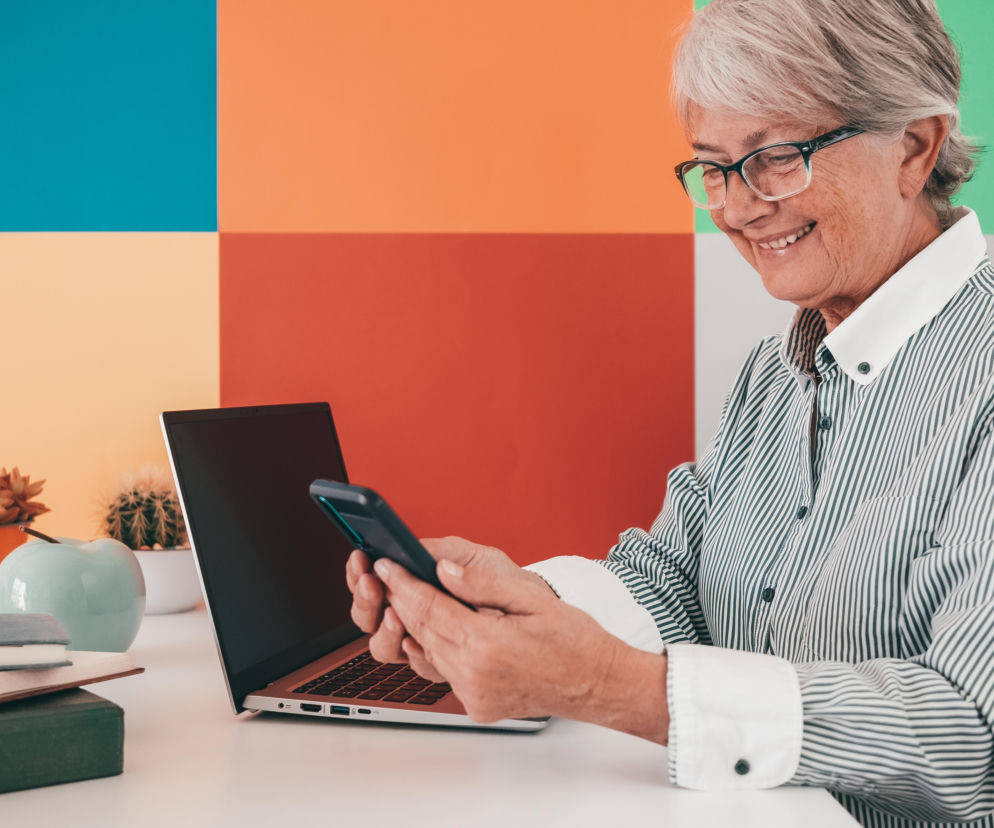 Uśmiechnięta seniorka w okularach korzysta ze smartfona przy laptopie. O zastrzeżeniu PESEL przeczytasz w SE Białystok.