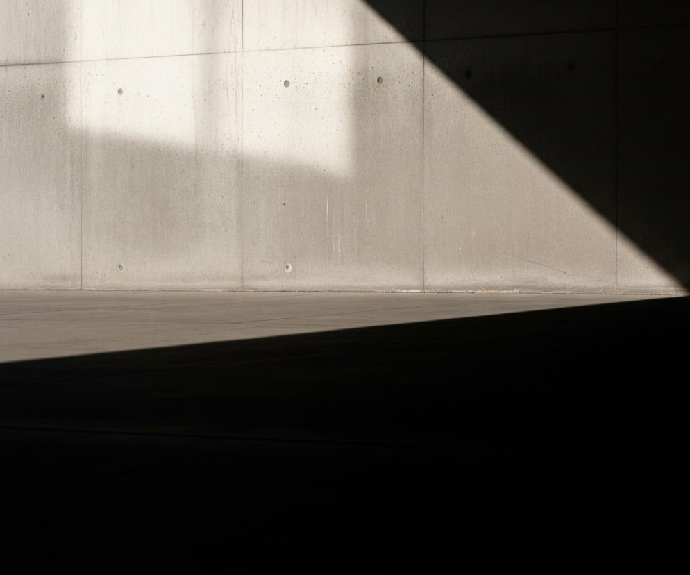 Jasnoszara betonowa ściana przecina kadr w pionie i poziomie, tworząc wyraźne linie na tle błękitnego nieba. Na tej ścianie widoczne są okrągłe otwory i delikatne zacieki. Duża, trójkątna plama cienia pokrywa prawą i dolną część ściany oraz podłogi, tworząc kontrast z oświetlonymi fragmentami. Na horyzoncie, po lewej stronie, rozmyte budynki z białymi elementami architektury majaczą w oddali.