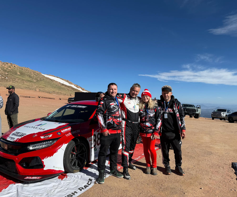 Maciej Serafin w kombinezonie rajdowym z zespołem przed czerwono-białym samochodem wyścigowym Honda Civic Type R na tle górzystego krajobrazu Pikes Peak. Zespół jest gotowy na wyścig, o czym można przeczytać więcej na naszym portalu.