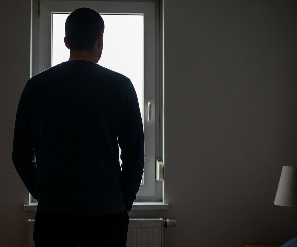 Mężczyzna stoi tyłem do obserwatora, patrząc w stronę jasnego okna, przez które wpada światło. Ma na sobie ciemną bluzę i spodnie, a jego sylwetka jest zarysowana przez kontrast z jasnym tłem okna. W lewym dolnym rogu okna widać biały grzejnik, a po prawej stronie obrazu znajduje się fragment łóżka z drewnianym zagłówkiem i granatową poduszką oraz lampa nocna z beżowym abażurem. Pomieszczenie jest słabo oświetlone, utrzymane w ciemnych, stonowanych barwach.