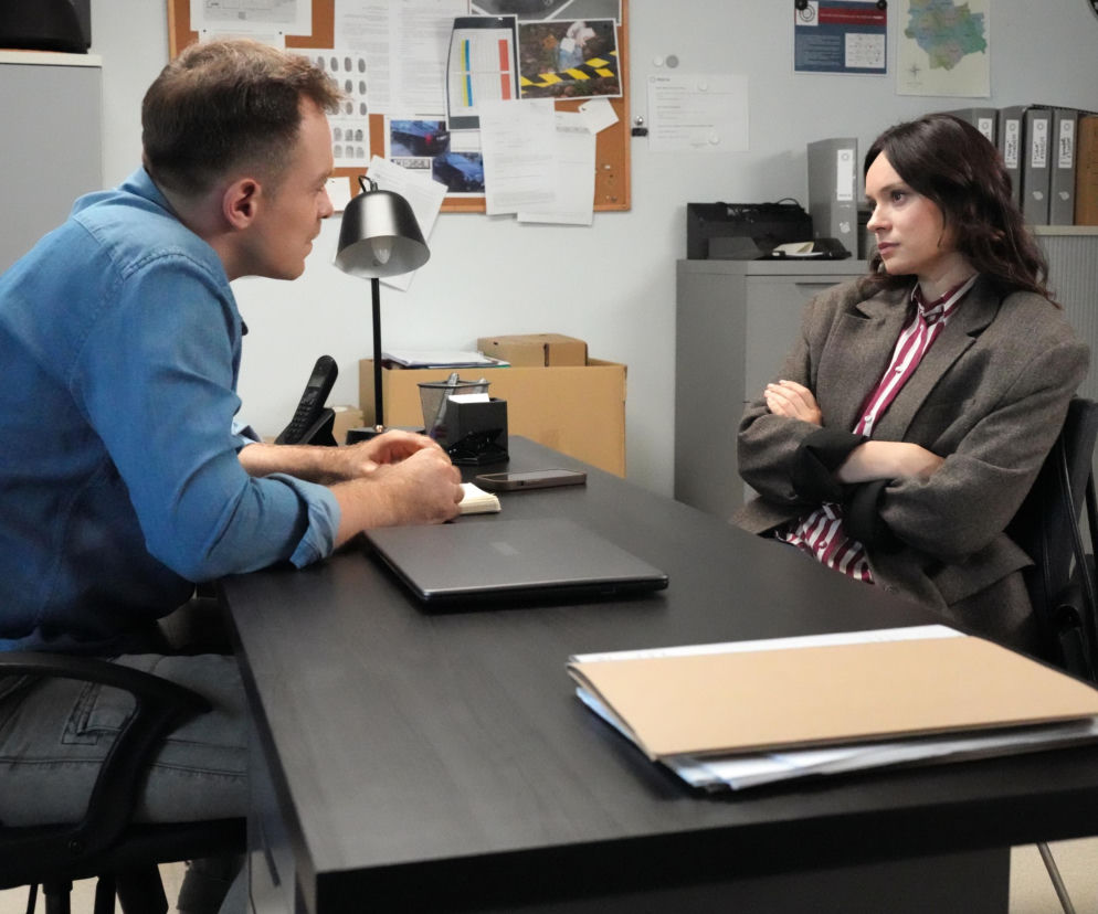 Młody mężczyzna w niebieskiej koszuli rozmawia z kobietą w marynarce. Siedzą naprzeciwko siebie przy biurku w biurze. Prawdopodobnie jest to kadr z serialu Barwy szczęścia, a więcej na temat serialu znajdziesz w Super Seriale.