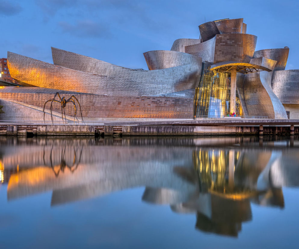 Nocny widok na Muzeum Guggenheima w Bilbao, którego tytanowa fasada, przypominająca pofalowane statki, odbija się w spokojnej wodzie rzeki Nervión. Przed budynkiem stoi monumentalna rzeźba pająka Maman Louise Bourgeois. To symbol rewitalizacji miasta, o czym można przeczytać na Architektura Murator Plus.