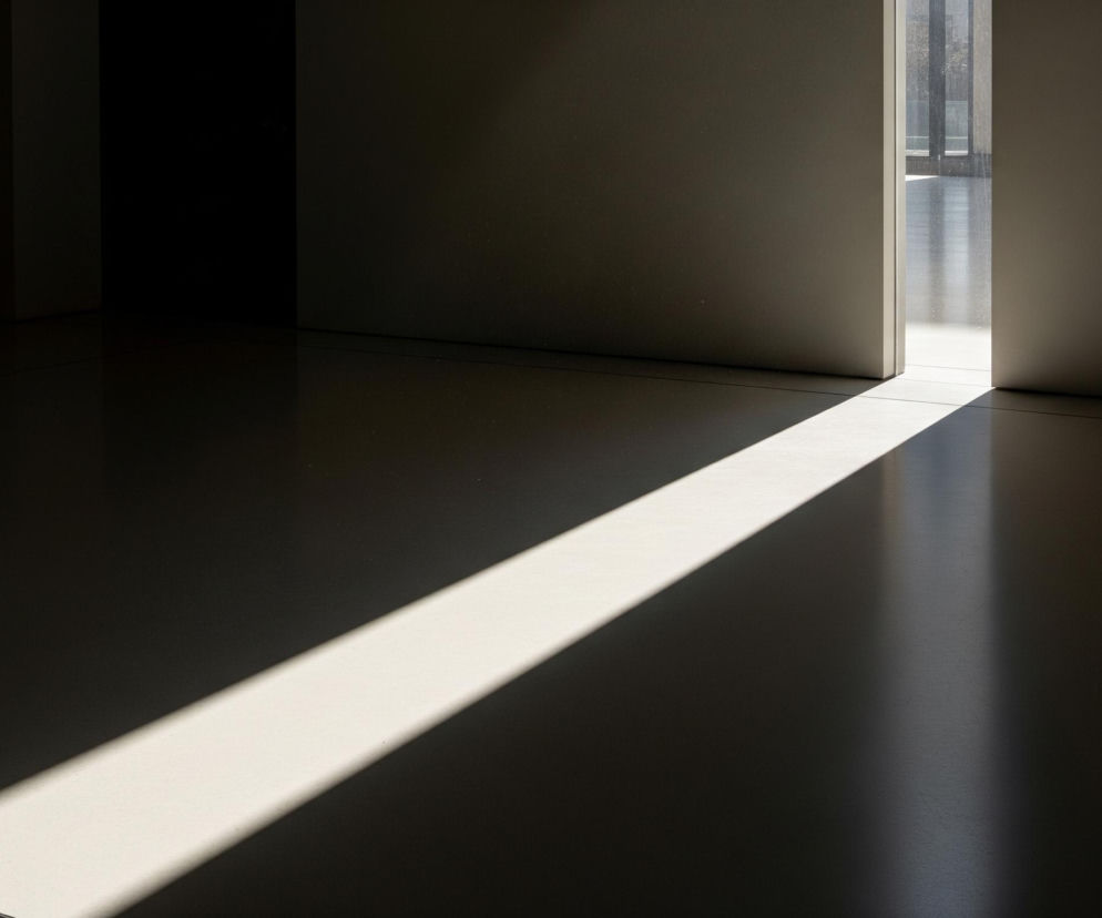 Wnętrze pomieszczenia z drzwiami lub przejściem po prawej stronie. Promień słońca wpada przez szczelinę drzwi, tworząc jasną, skośną linię na ciemnej podłodze, biegnącą od prawego górnego rogu w stronę lewego dolnego. Ściany są jasnobrązowe lub szare, a obszar po lewej stronie jest znacznie ciemniejszy, pogrążony w cieniu. Za otwartymi drzwiami widać zarys zewnętrznego otoczenia, przypominający okna lub przeszkloną ścianę, z widocznymi refleksami światła.