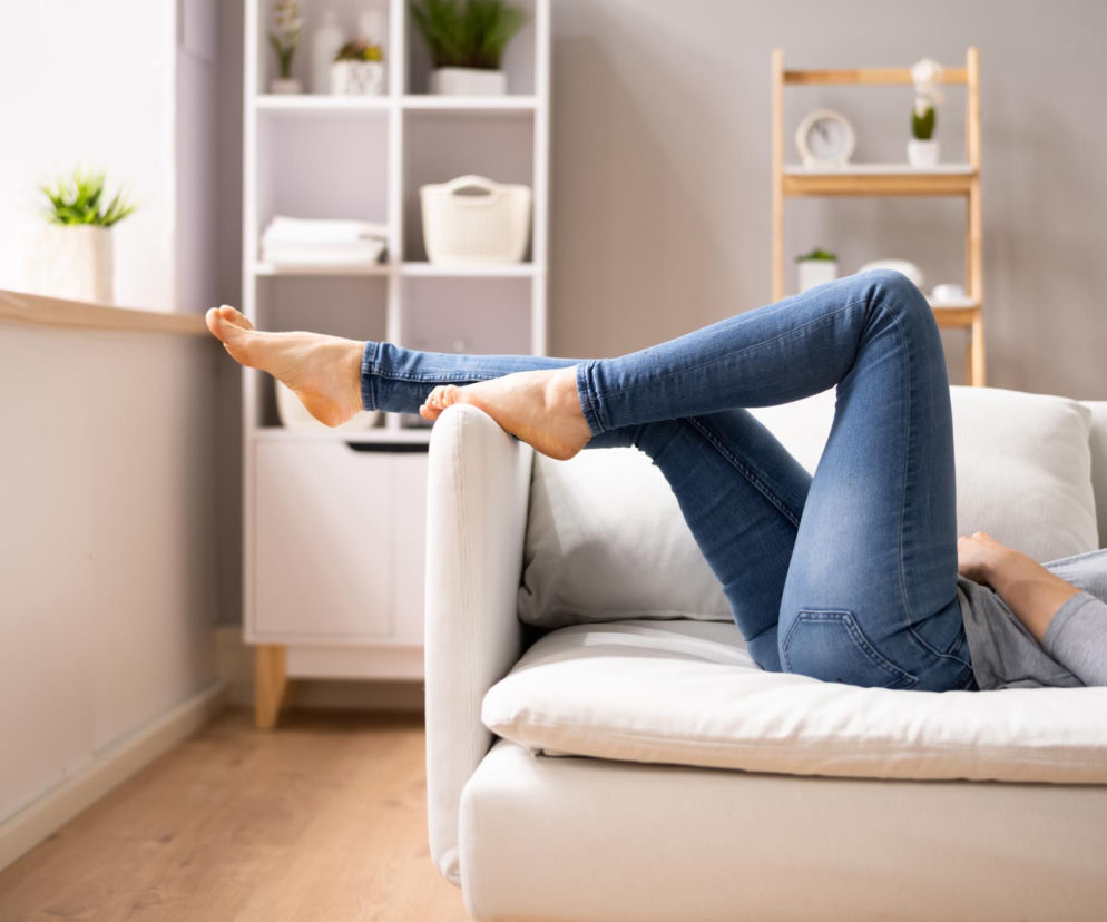 Kobieta odpoczywająca na białej kanapie z uniesionymi nogami, ubrana w dżinsy i szarą bluzkę. Sposób na ulgę dla opuchniętych i ciężkich nóg, o czym przeczytasz więcej na Poradnik Zdrowie.