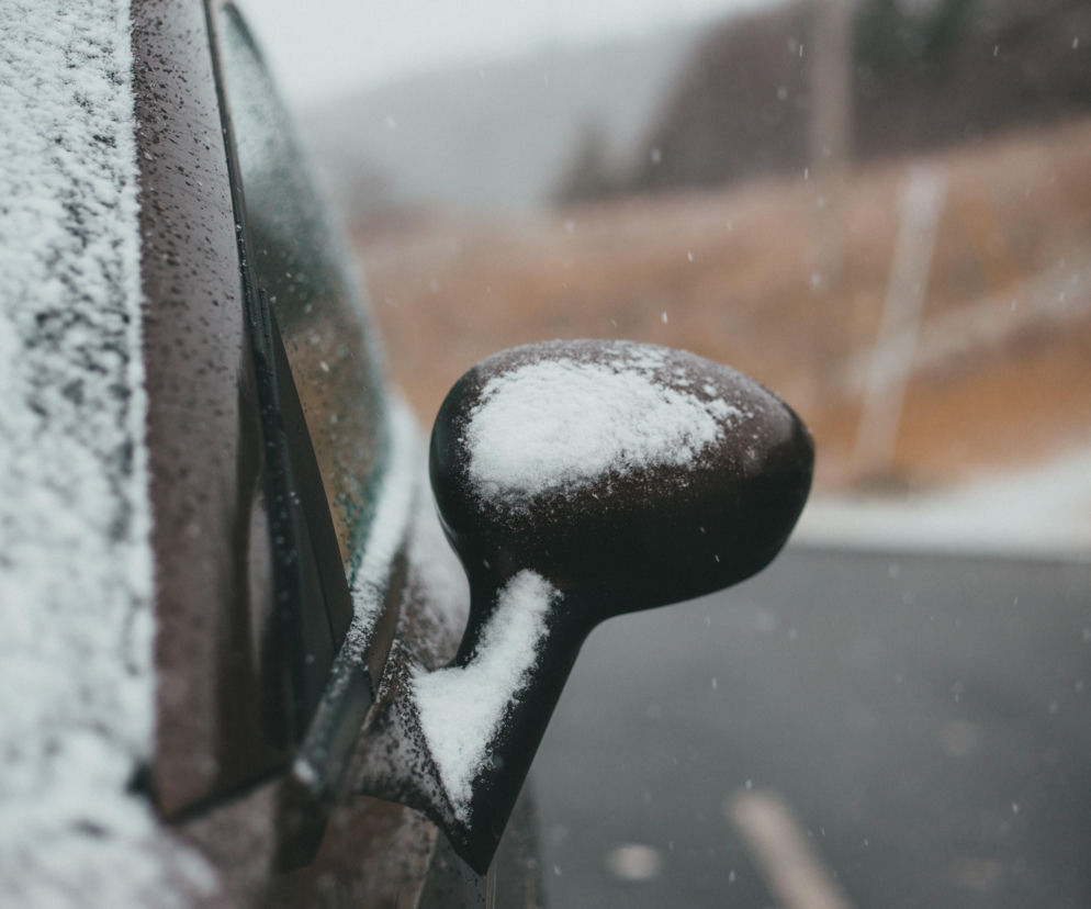 Lusterko boczne samochodu pokryte śniegiem, symbolizujące zimowe warunki na drogach. Obraz nawiązuje do kar za nieodśnieżone auta. Więcej o mandatach za jazdę w mrozy przeczytasz na Super Biznes.