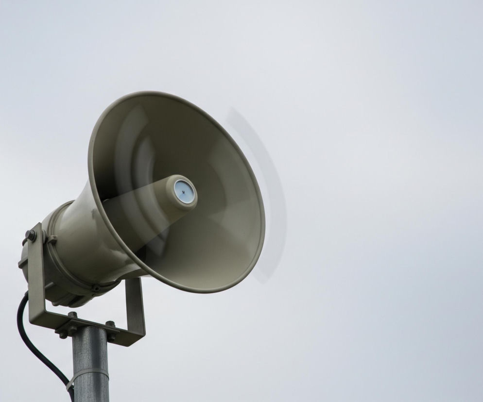 Pojedynczy głośnik tubowy w odcieniu khaki, zamontowany na metalowym słupie po lewej stronie kadru. Głośnik skierowany jest w prawo, a jego wnętrze ma jaśniejszy odcień, z centralnym elementem przypominającym głośnik w kolorze jasnoniebieskim lub białym. Dookoła tego centralnego elementu widoczne są rozmyte, poziome linie, sugerujące wibracje lub ruch dźwięku. Głośnik przymocowany jest do słupa za pomocą ciemnego, metalowego wspornika i kilku śrub, a od spodu biegnie gruby, czarny kabel. Tło stanowi jednolite, jasnoszare niebo, bez widocznych chmur czy innych elementów.