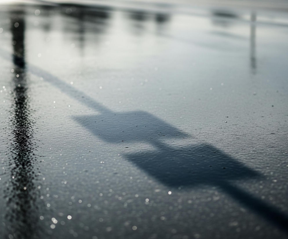 Na pierwszym planie widoczna jest mokra, ciemna nawierzchnia pokryta licznymi drobnymi kroplami wody, które odbijają światło, tworząc efekt bokeh. Przez środek obrazu, od lewej strony, przebiega ciemny, pionowy cień słupa, rozciągający się do dolnej krawędzi. W jego prawą stronę odchodzi cień poziomy, tworząc kształt litery T lub strzałki skierowanej w prawo, zanikający w oddali. Tło jest rozmyte, z niewyraźnymi odcieniami szarości i bieli, sugerującymi odbicia drzew lub innych obiektów w lśniącej powierzchni.