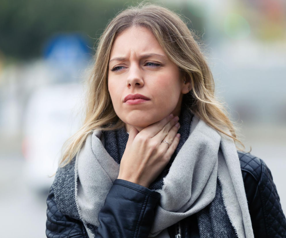 Młoda kobieta z jasnymi włosami, w szarym szaliku i czarnej ramonesce, masuje szyję, wyrażając dyskomfort związany z bólem gardła lub chrypką. Na Poradnik Zdrowie znajdziesz sposoby na cichy refluks, który może być przyczyną tych dolegliwości.