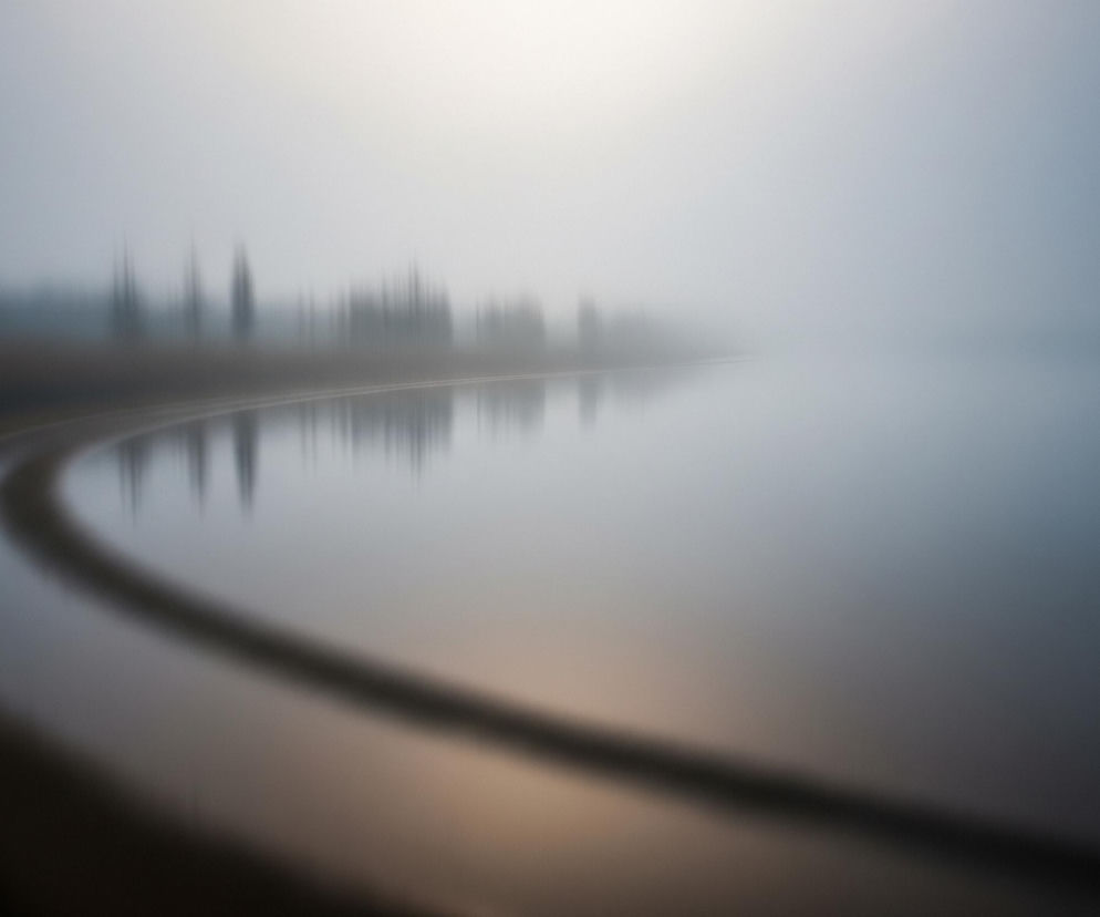 Mglisty krajobraz z akwenem wodnym i odległą linią drzew, rozmyty i stylizowany. Po lewej stronie widoczny jest ciemny brzeg lub pas lądu, który łagodnie zakrzywia się ku górze, a następnie w prawo, tworząc wyraźny łuk. Nad brzegiem, w oddali, rysują się niewyraźne, pionowe kształty drzew, zanurzone w gęstej, szaro-niebieskiej mgle. Odbicia drzew i światła w wodzie są zamazane, co sugeruje ruch lub specjalny efekt fotograficzny, dodając obrazowi eterycznego charakteru. Całość utrzymana jest w chłodnej palecie barw, z dominacją odcieni szarości, błękitu i delikatnego, rozświetlonego beżu na horyzoncie.