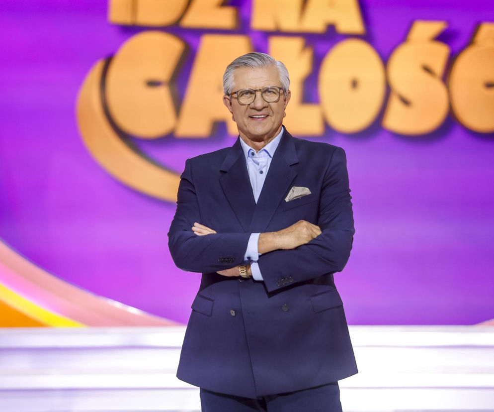 Uśmiechnięty Zygmunt Chajzer w garniturze na tle logo programu Idź na całość. O kulisach show przeczytasz na SE.