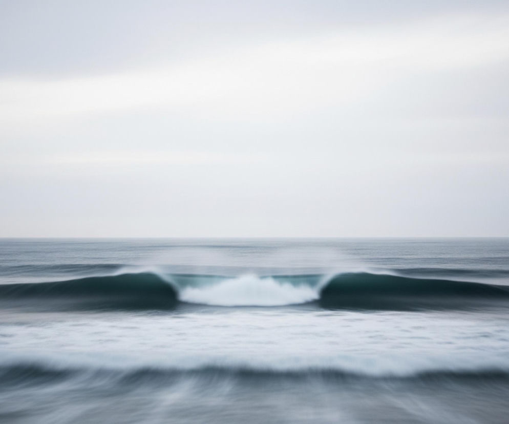 Pośrodku kadru znajduje się duża fala morska, której centralna część załamuje się, tworząc tunel z białą, rozpyloną pianą w środku. Boczne części fali są gładkie i ciemnoniebiesko-zielone, przechodząc w mniejsze, rozmyte fale na pierwszym planie, gdzie powierzchnia wody jest pofałdowana i lekko zamglona. Horyzont oddziela spokojne, ciemnoszare morze od jednolitego, jasnoszarego nieba, pokrytego cienkimi warstwami chmur.