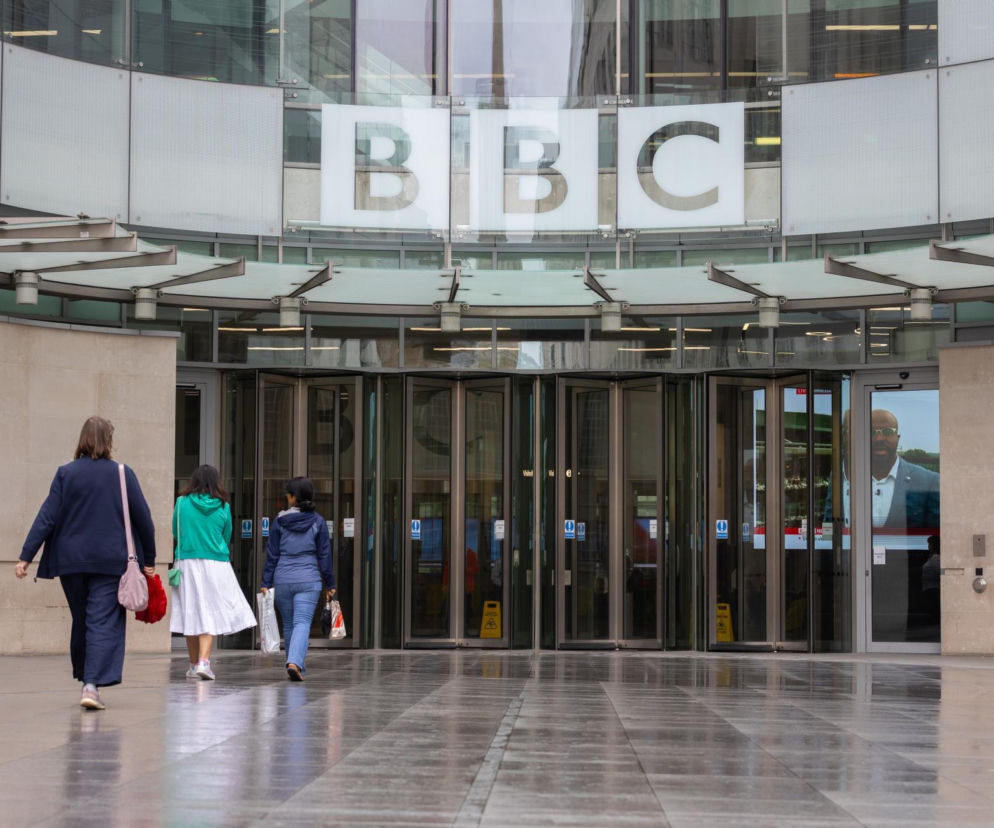 Wejście do budynku BBC z dużym, białym logo stacji i przeszklonymi drzwiami obrotowymi. Przed wejściem widoczne są sylwetki ludzi, co odzwierciedla sytuację masowych zwolnień w firmie medialnej, o których można przeczytać na Super Biznes.