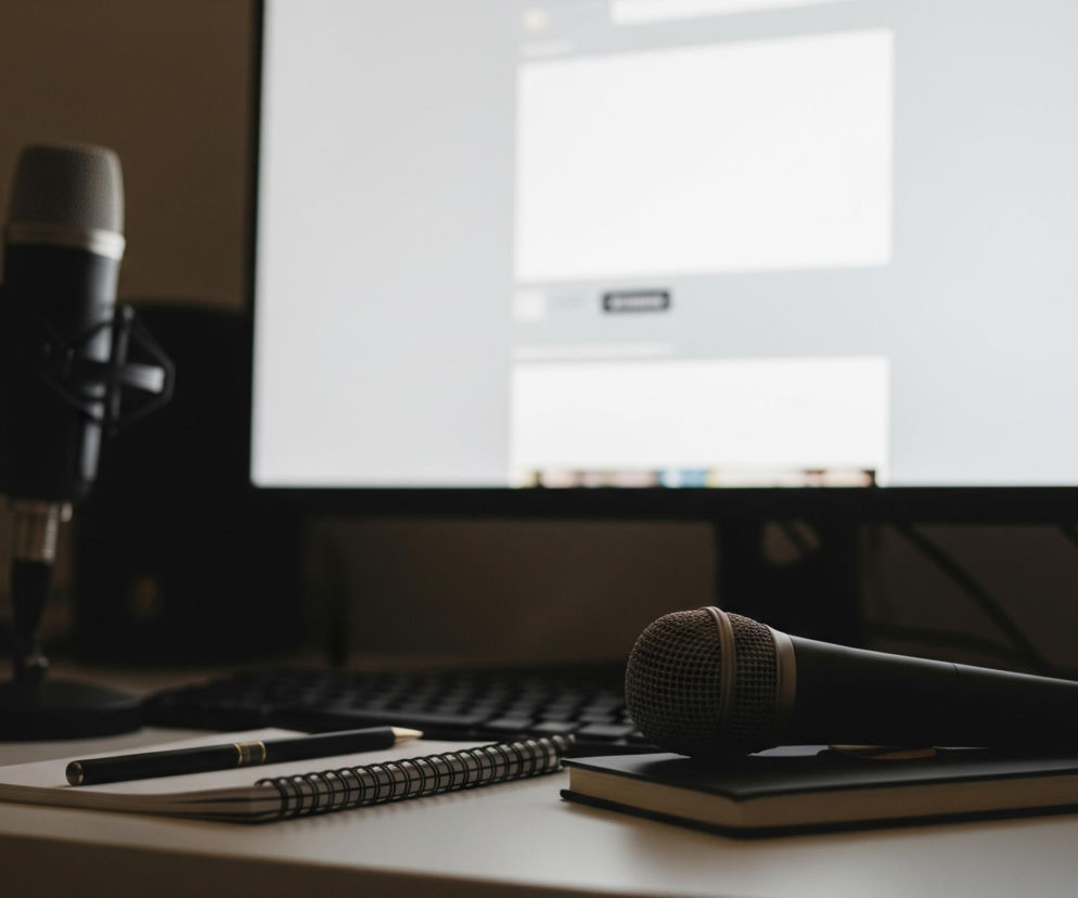 Na pierwszym planie, po prawej stronie leży czarny mikrofon z szarą siatką na głowicy, oparty na małym, czarnym notesie. Obok, po lewej, znajduje się otwarty zeszyt na spirali, a na nim czarny długopis z metalowym akcentem. W tle widoczny jest monitor komputera z jasnym ekranem, wyświetlającym niezidentyfikowaną treścią o strukturze stron internetowych z białymi, pustymi polami i ciemnymi paskami. Po lewej stronie ekranu monitora znajduje się drugi mikrofon, stojący pionowo na statywie, niewyraźny i częściowo zasłonięty, a dalej, w tle, widoczny jest zarys kolejnego ciemnego monitora lub innego urządzenia. Cała scena jest utrzymana w ciemnej tonacji, sugerującej słabe oświetlenie pomieszczenia.