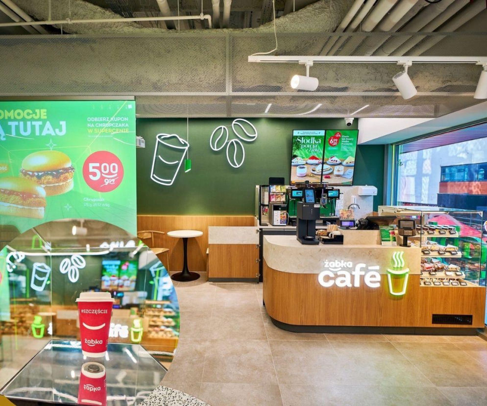 Wnętrze Żabka Cafe z ladą i strefą kawową. Na miniaturze czerwony kubek Żabki. O 13 000. sklepie przeczytasz na Eska.