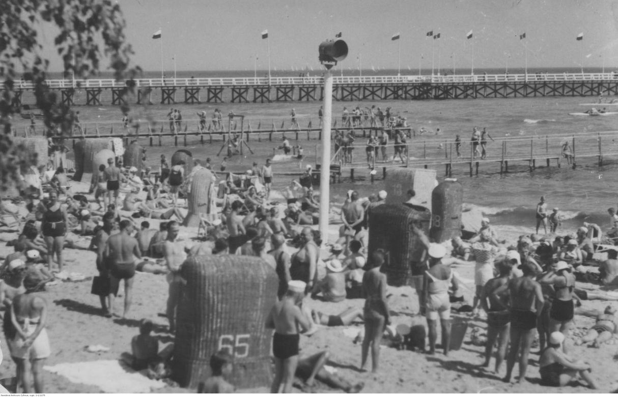 Plażowicze podczas wypoczynku na morzem Bałtyckim. Widoczne wydzielone kąpielisko, w tle molo. Zdjęcie zrobione w okresie 1918-1939