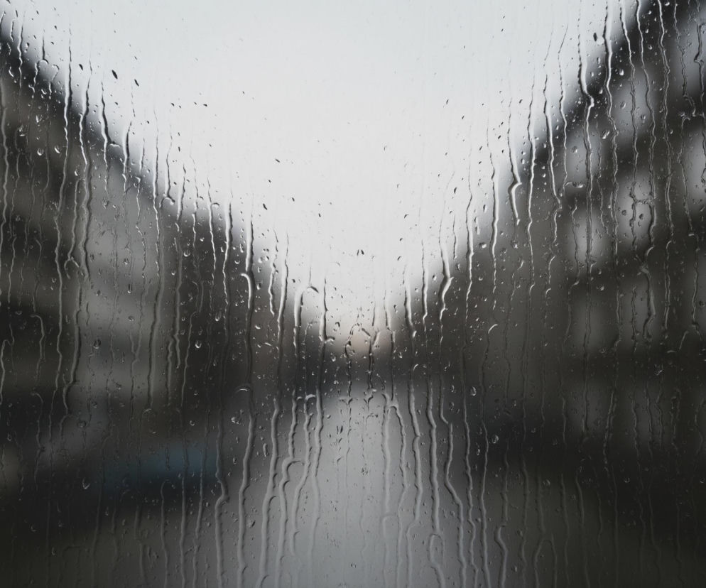 Przednią część obrazu wypełnia szyba pokryta licznymi kroplami deszczu i smugami wody spływającymi pionowo. W tle, rozmyte i nieostre, widoczne są kontury budynków o ciemnoszarych i brązowych odcieniach, zlewające się z jasnoszarym, niemal białym, niebem w górnej części kadru. Pomiędzy budynkami, w centralnej dolnej części, zarysowuje się wąska, ciemniejsza przestrzeń, sugerująca ulicę lub drogę.