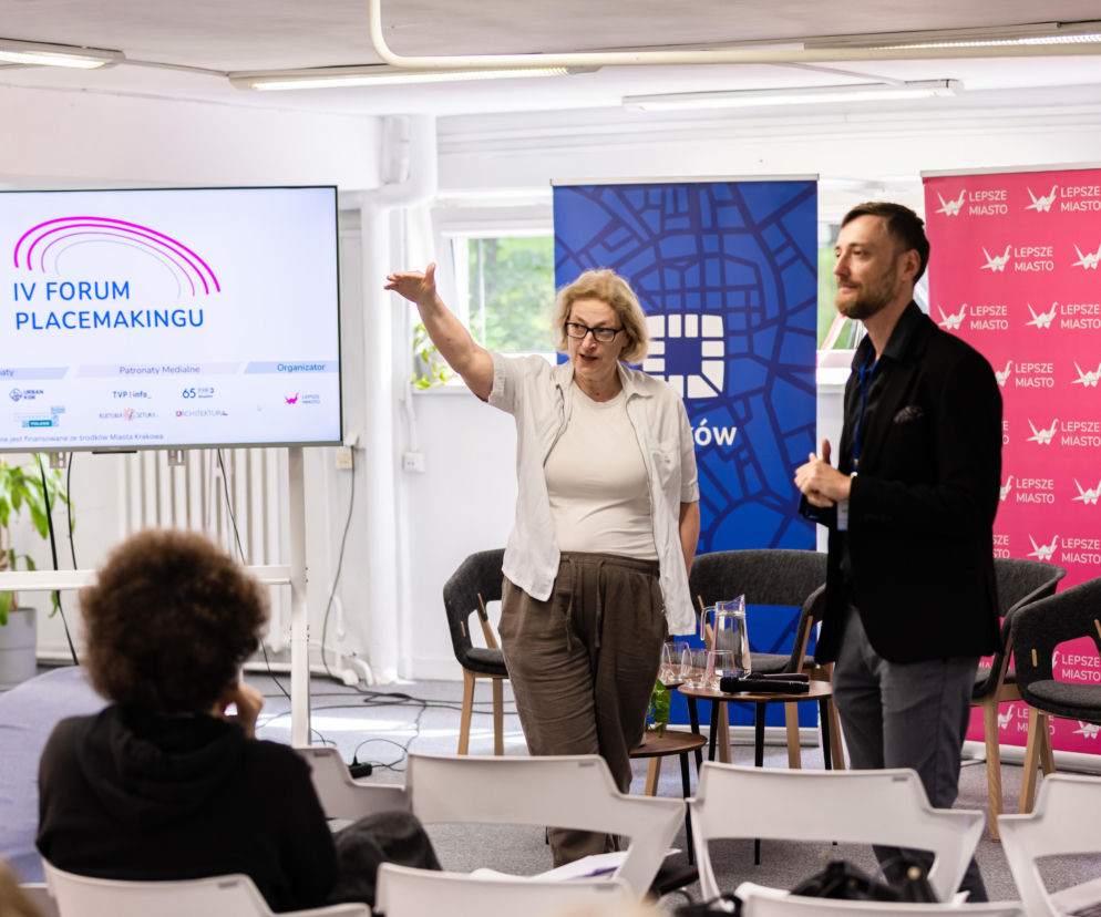 Prezentacja „IV Forum Placemakingu” na ekranie podczas wydarzenia, gdzie Carolina Pietyra, dyrektorka Krakowskiego Biura Festiwalowego, gestykuluje, a obok stoi mężczyzna. Na ścianie flagi „Lepsze Miasto”. O temacie tym przeczytasz więcej na portalu Architektura Murator Plus.