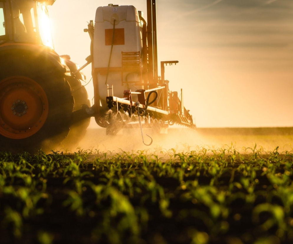 Ciągnik rolniczy z opryskiwaczem na polu, spryskujący młode uprawy podczas wschodu słońca. Rolnicy mogą przeczytać o zagrożeniach związanych z pyłem podczas oprysków i sprawdzić skuteczność masek i kabin na portalu Super Biznes.