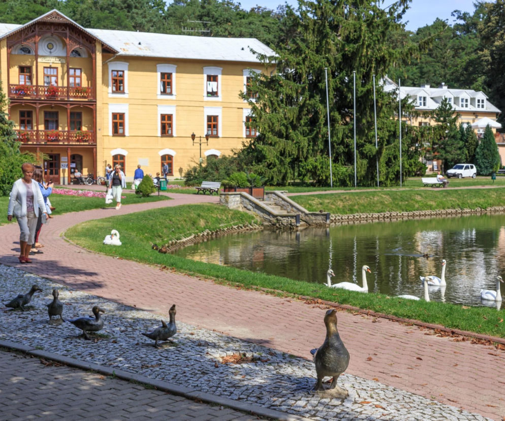 Sanatorium z żółtą fasadą i drewnianymi zdobieniami, w pobliżu jeziora z łabędziami i rzeźbami kaczek. Ludzie spacerują po alejkach. Zdjęcie ilustruje temat artykułu o zwrotach miejsc w sanatoriach, dostępnych na Super Biznes.