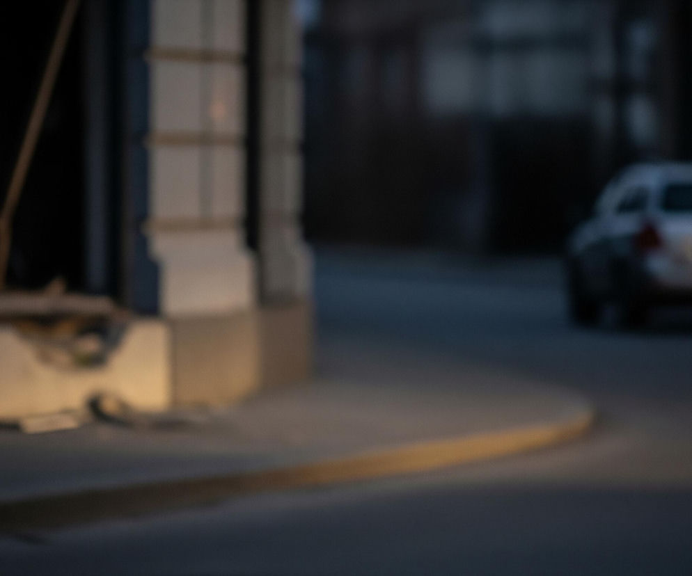 Lewa strona zdjęcia przedstawia fragment jasnego, kremowego budynku z widocznymi poziomymi i pionowymi elementami architektonicznymi, przypominającymi kolumny lub pilastry. U dołu fasady widoczne jest ciepłe, żółte światło, rozjaśniające chodnik tuż przy krawędzi budynku. W centrum obrazu znajduje się ciemnoszary chodnik, łagodnie przechodzący w ulicę, a w tle, po prawej stronie, jedzie srebrne kombi z wyraźnie widocznym, okrągłym światłem tylnej lampy. Całe tło, poza budynkiem i samochodem, jest rozmazane i utrzymane w ciemnych, niebieskawo-szarych tonacjach, sugerujących zmierzch lub noc.