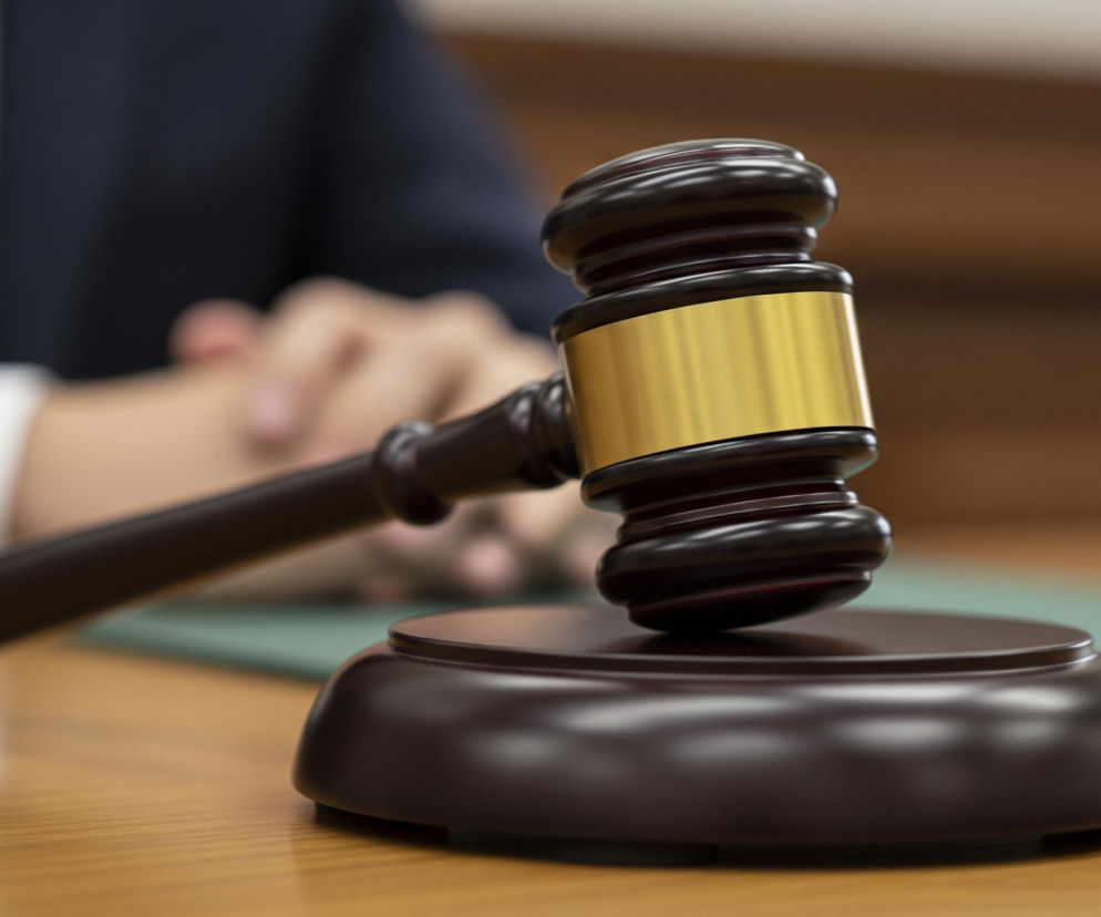 Młotek sędziowski wykonany z ciemnego drewna z metalową, złotą opaską na głowicy leży na dopasowanej, okrągłej podstawie. Przedmioty te spoczywają na jasnym, drewnianym biurku, za którym siedzi rozmyta postać w granatowym garniturze z niebieskim krawatem i białą koszulą, z dłońmi splecionymi przed sobą. W tle, na biurku widoczna jest zielona podkładka, a dalej jasnobrązowa ściana.