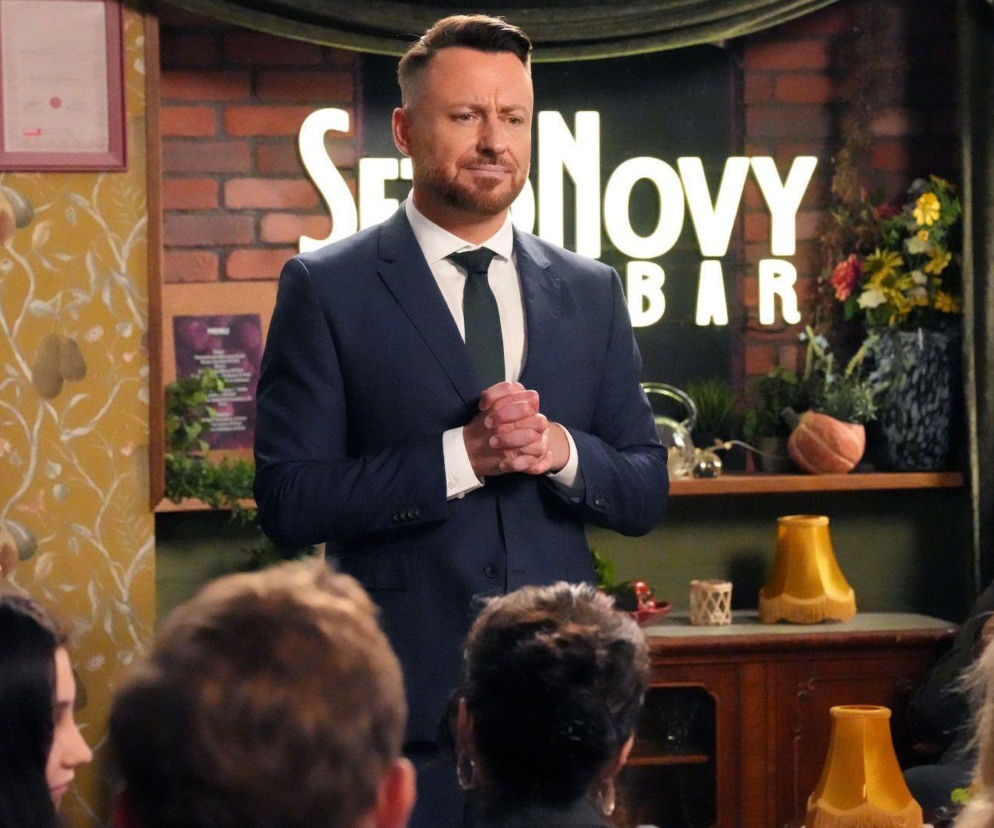 Adam Szczyszczaj jako Wilk w granatowym garniturze z zielonym krawatem, stojący przed tablicą z napisem S Novy BAR, ze złożonymi dłońmi, rozmawia z grupą mieszkańców. Scena nawiązuje do serialu Barwy szczęścia, o którym więcej przeczytasz na portalu Super Seriale.