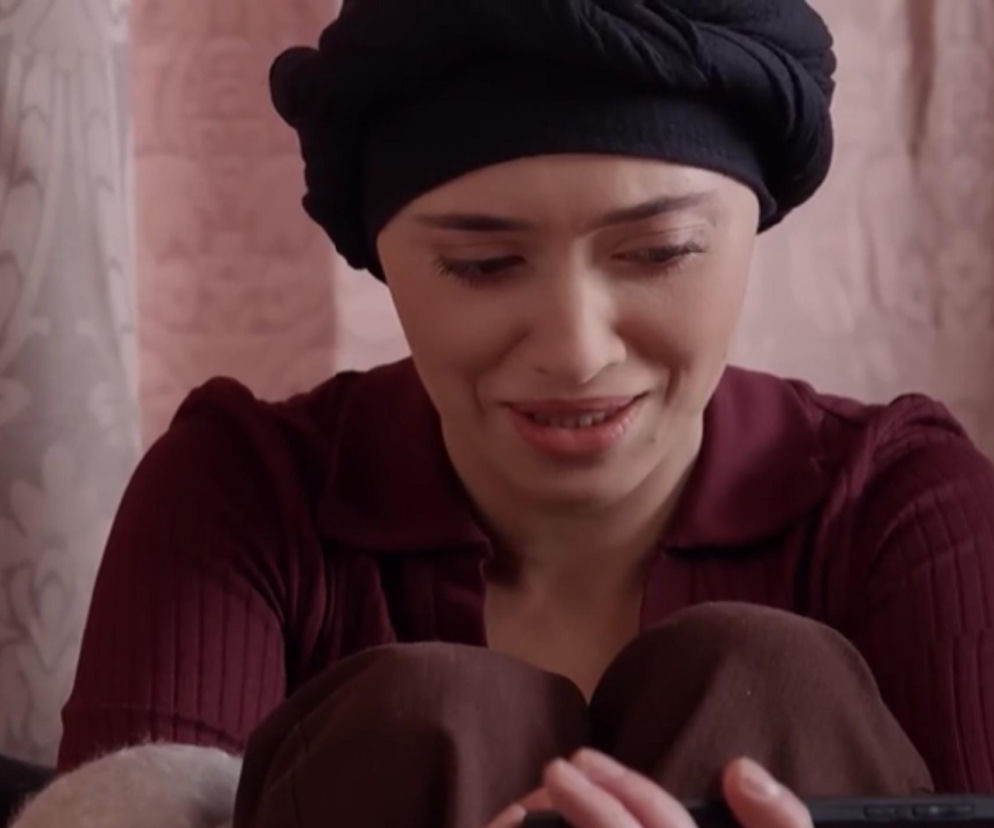 Aktorka Patrycja Franczak jako Angelika w scenie z serialu Pierwsza miłość, siedząca, ubrana w burgundową bluzkę i czarny turban, z delikatnym uśmiechem na twarzy, trzymająca coś w dłoniach, co może zwiastować nowe wyzwania w serialu opisanym na naszym portalu.