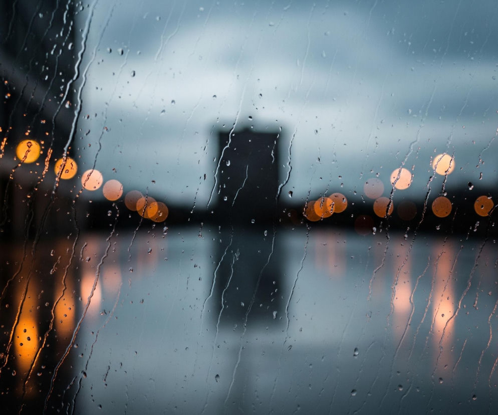 Na pierwszym planie widoczna jest mokra szyba pokryta licznymi kroplami wody oraz śladami spływającej deszczówki, tworzącymi pionowe smugi. Przez zamazane szkło rozciąga się krajobraz miejski, z rozmytymi, pomarańczowymi światłami bokeh, które odbijają się w lustrzanej powierzchni poniżej. W centralnej części tła, w oddali, rysuje się ciemna, prostokątna sylwetka budynku, a horyzont zamyka szare, zachmurzone niebo.