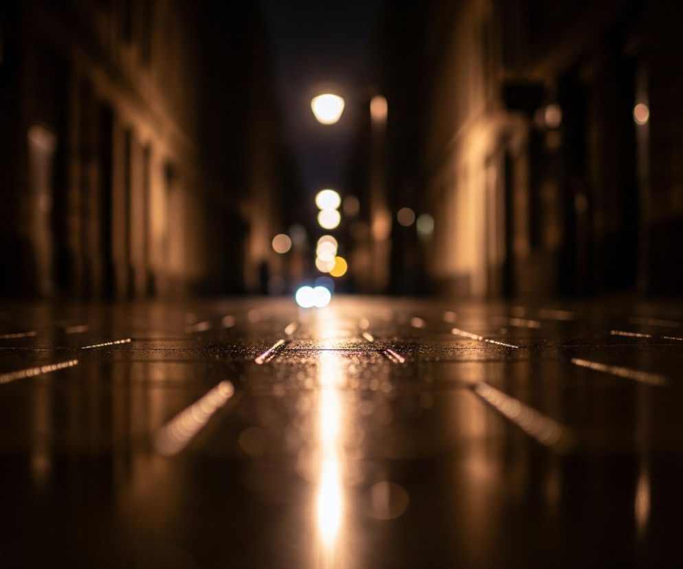 Widoczna jest wąska ulica nocą, oświetlona ciepłym światłem latarni. Chodnik i jezdnia, zajmujące dolną połowę kadru, są mokre, co tworzy długie, lśniące odbicia świateł. W głębi ulicy widać pojedynczą, jaskrawą lampę uliczną oraz rozmyte, punktowe światła o różnej intensywności, w tym parę jasnych, niebieskawo-białych świateł pojazdu. Po obu stronach ulicy ciągną się zamazane fasady budynków, zabarwione na odcienie brązu i pomarańczu przez padające światło.